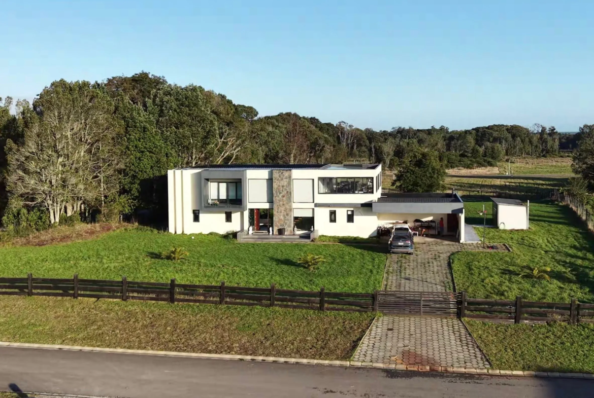 Casa de dos pisos rodeada de bosque nativo, con camino de acceso empedrado y jardín